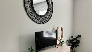 Un salon moderne avec un téléviseur à écran plat posé sur un meuble en bois clair, un miroir décoratif et deux plantes en pot.