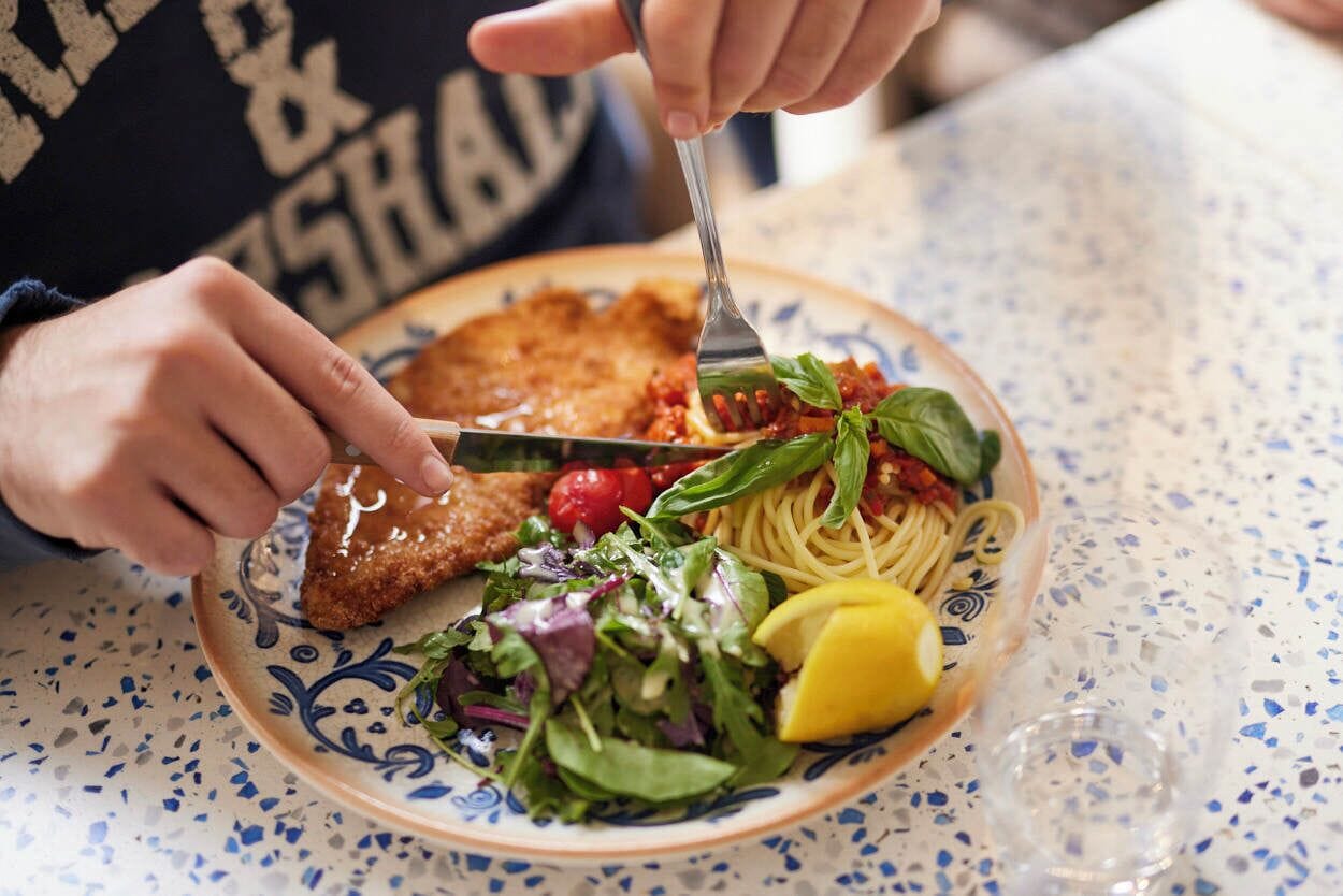 Una persona cortando una milanesa en un plato con espagueti, ensalada y un gajo de limón.