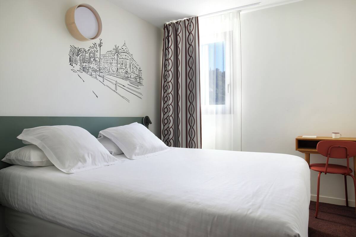 Un lit bien fait, avec du linge de lit blanc, un bureau avec une chaise et un mur orné d’une fresque représentant un paysage urbain en noir et blanc.
