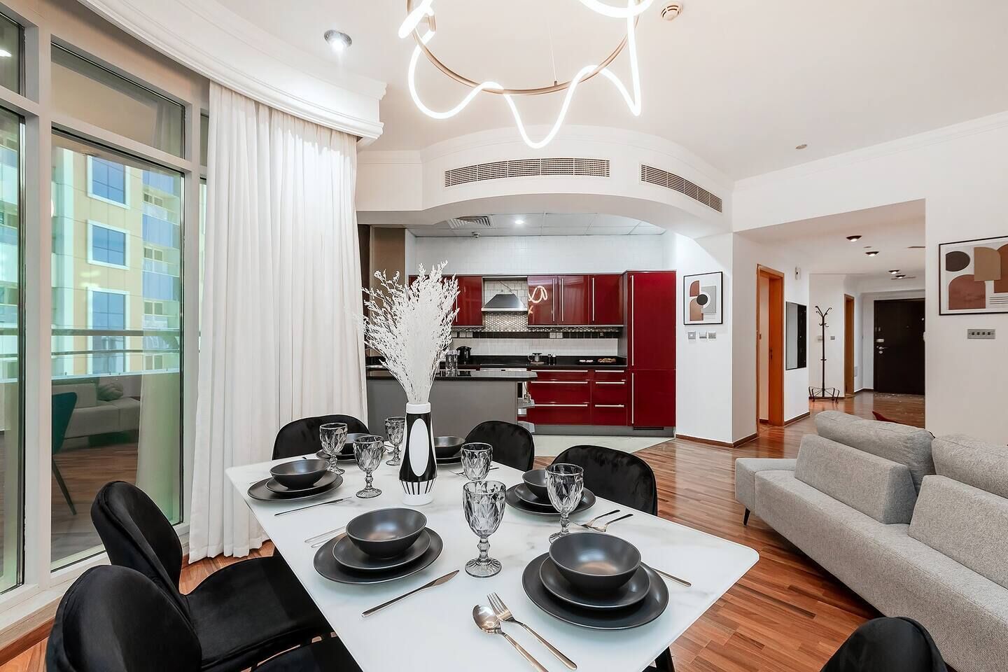 A modern dining area with a white table set for six, a kitchen in the background, and a view of the city through large windows.