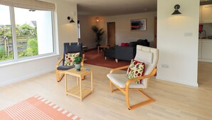 A modern living room with a sofa, two armchairs, a small table, and a view of the garden through large windows. - Pembroke (Grange-over-Sands)