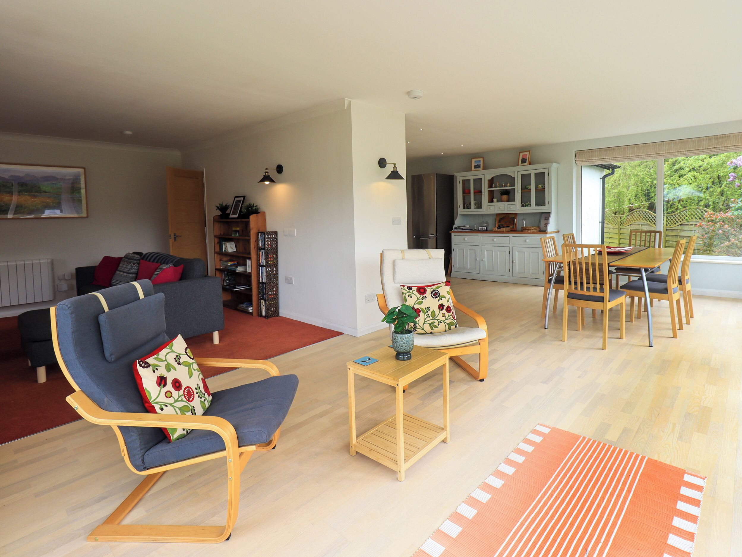 A spacious living area with a sofa, a dining table with chairs, a kitchen in the background, and a view of greenery outside.