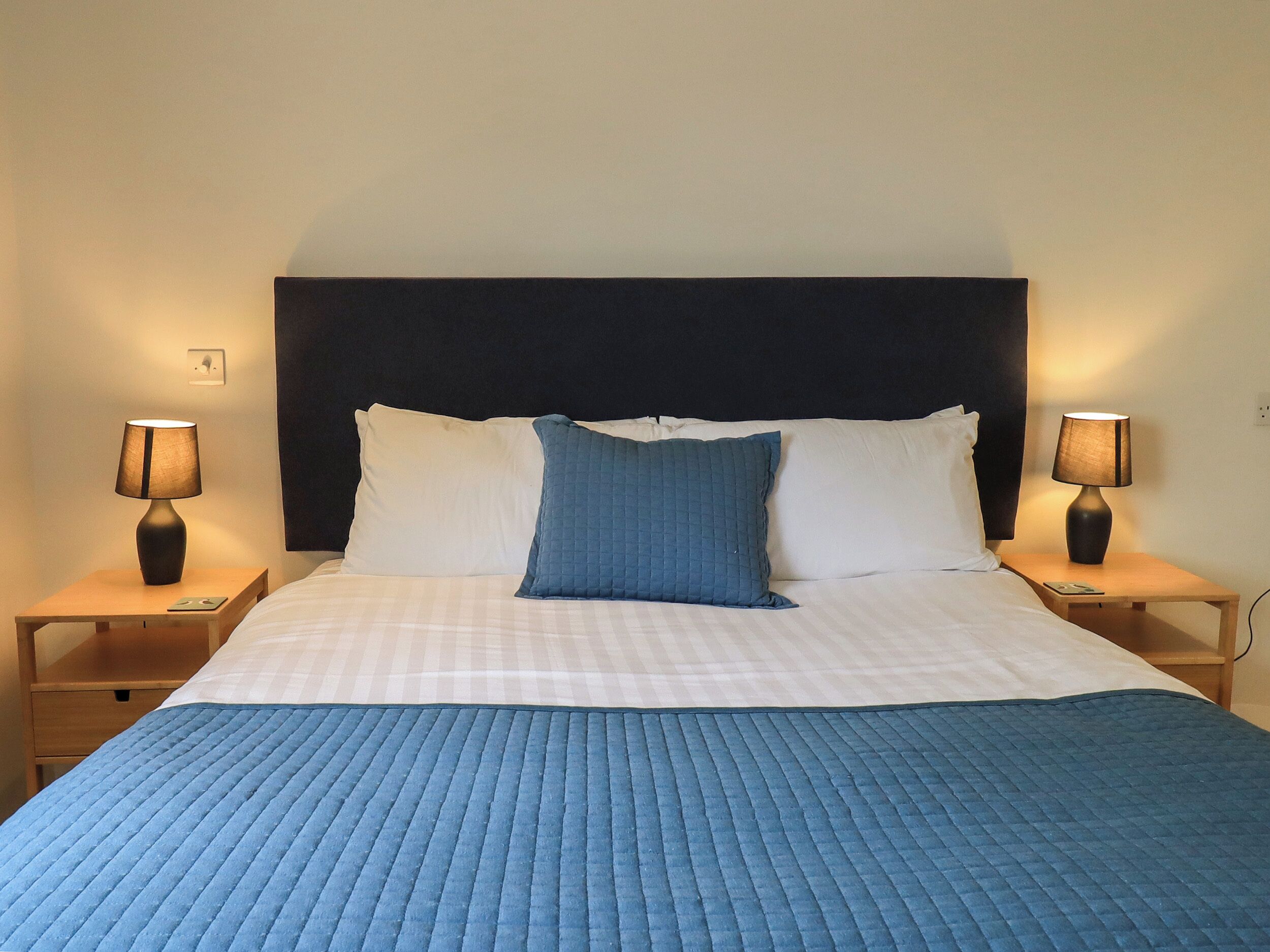 A neatly made bed with a blue quilt, two bedside lamps, and a dark headboard.