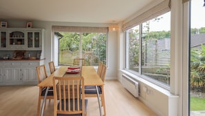 A dining area with a wooden table and chairs, a buffet cabinet, and large windows with a view of a garden. - Pembroke (Grange-over-Sands)