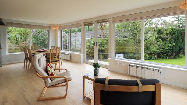 A spacious living room with a dining area, large windows, and a view of a garden. - Pembroke (Grange-over-Sands)
