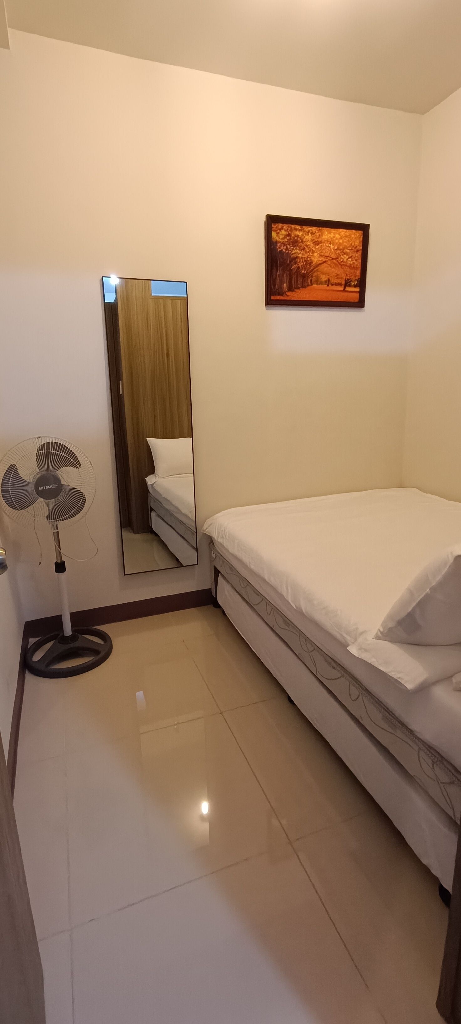 Habitación de hotel con una cama, un ventilador de pie, un espejo y un cuadro en la pared.