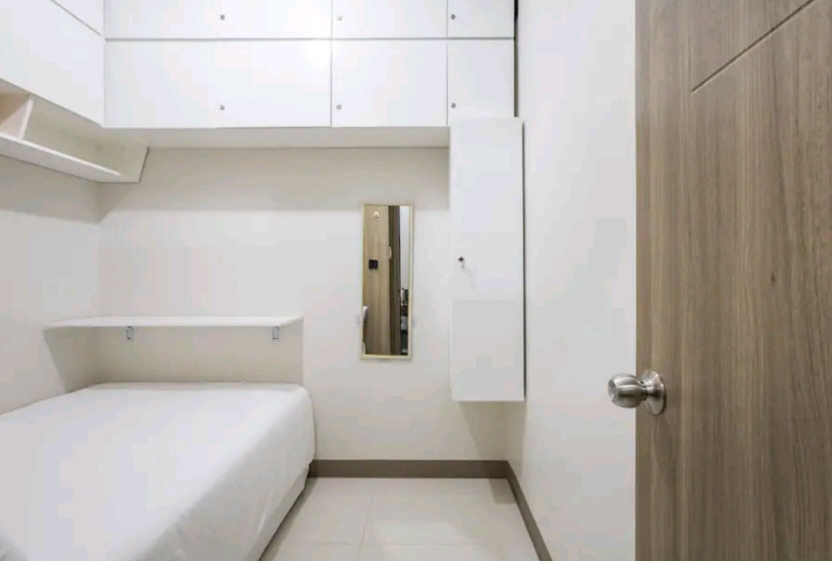 A small, compact room with a bed, a wooden door, and white cabinets.