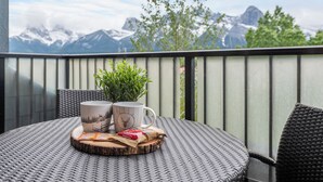 A round table with two mugs, a plant, and a packet of snacks on a woven tablecloth, overlooking a mountainous landscape. - Heavenly Mountain Condo - Outdoor Hot Tubs & Steam Room (Canmore)