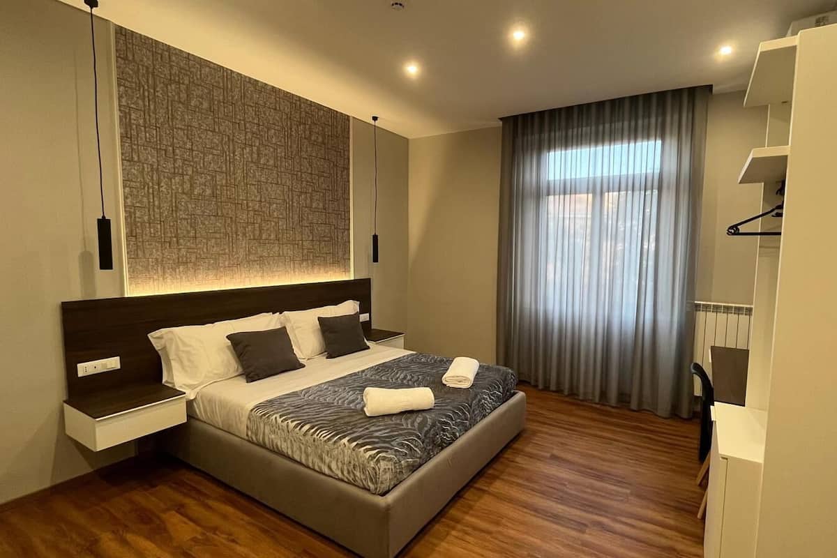 A modern bedroom with a large bed, a nightstand, a wardrobe, and a window with curtains.