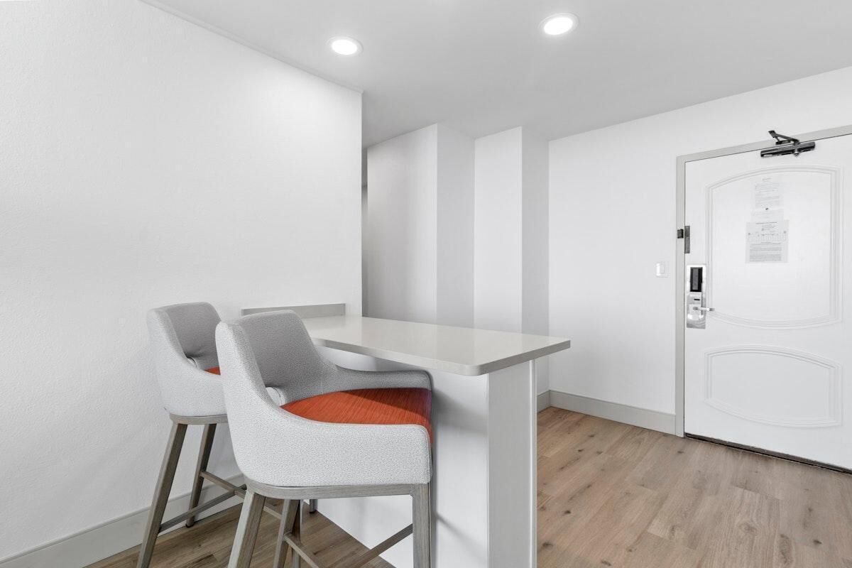 Una stanza moderna e minimalista con un tavolo bianco e due sgabelli da bar, una porta bianca con maniglia e un soffitto con illuminazione a incasso.
