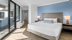 Une chambre d’hôtel moderne avec un grand lit, deux tables de chevet avec des lampes, une vue sur un balcon extérieur accessible par une porte vitrée, et un mur d’accent bleu.