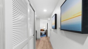 A hallway with a wooden floor, white walls, and a door with horizontal blinds. There are framed pictures on the wall and a bed with a blue and white striped blanket in the background.