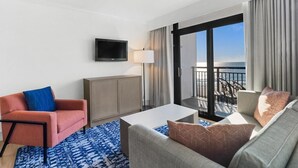 A hotel room with a sofa, armchair, flat-screen TV, and a balcony with a view of the ocean.