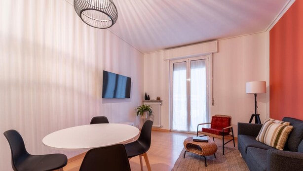 A modern living room with a round dining table, black chairs, a grey sofa, and a red armchair. There is a TV mounted on the wall, a floor lamp, and a window with curtains. - Appartamento Gelso by Wonderful Italy (Como)