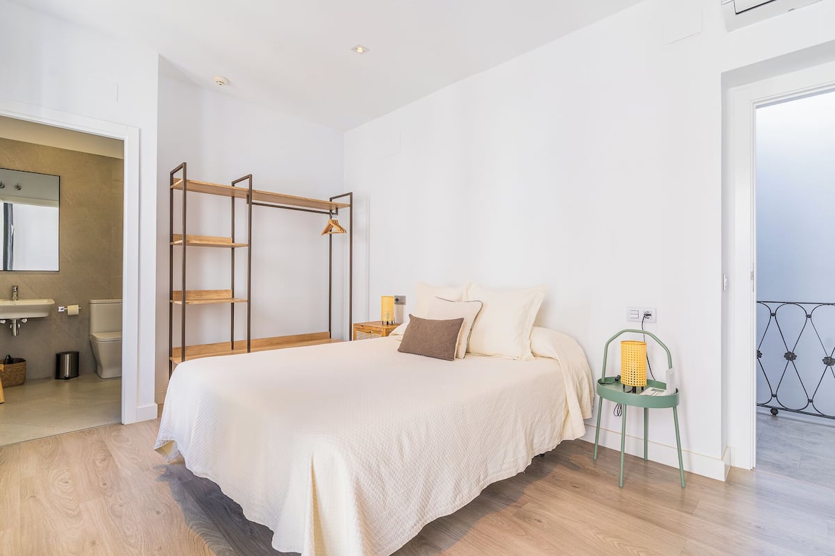 A bedroom with a bed, a wooden shelf, a bedside table with a lamp, and a view of the balcony.