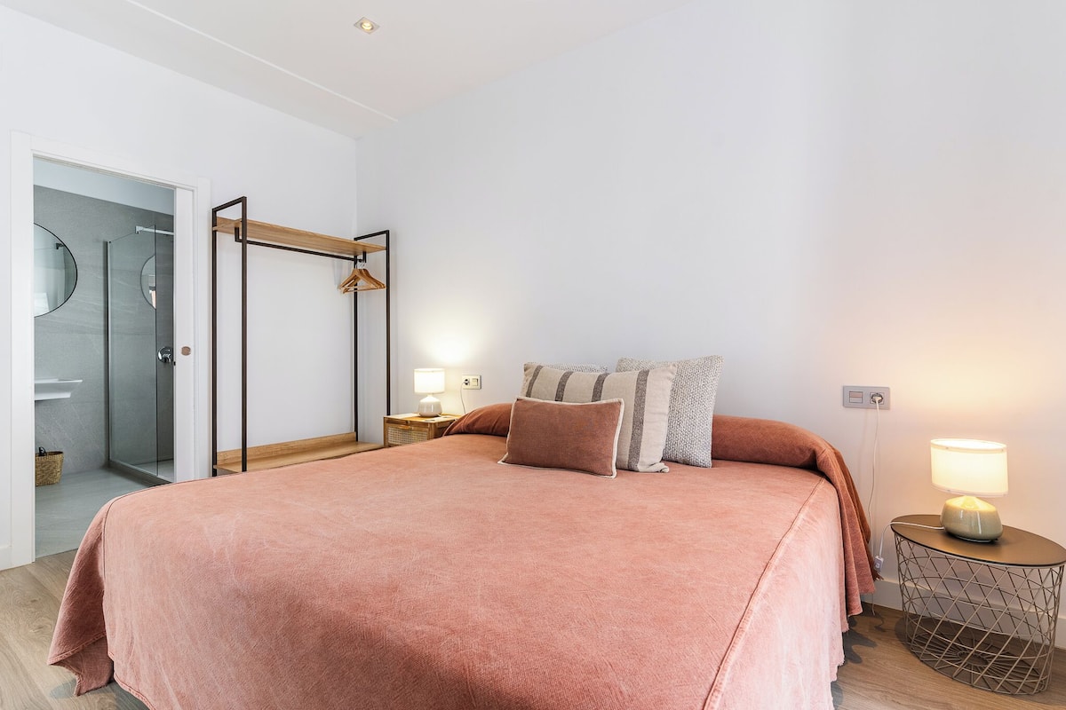 A bedroom with a large bed, a bedside table with a lamp, a wardrobe, and a glass shower area.