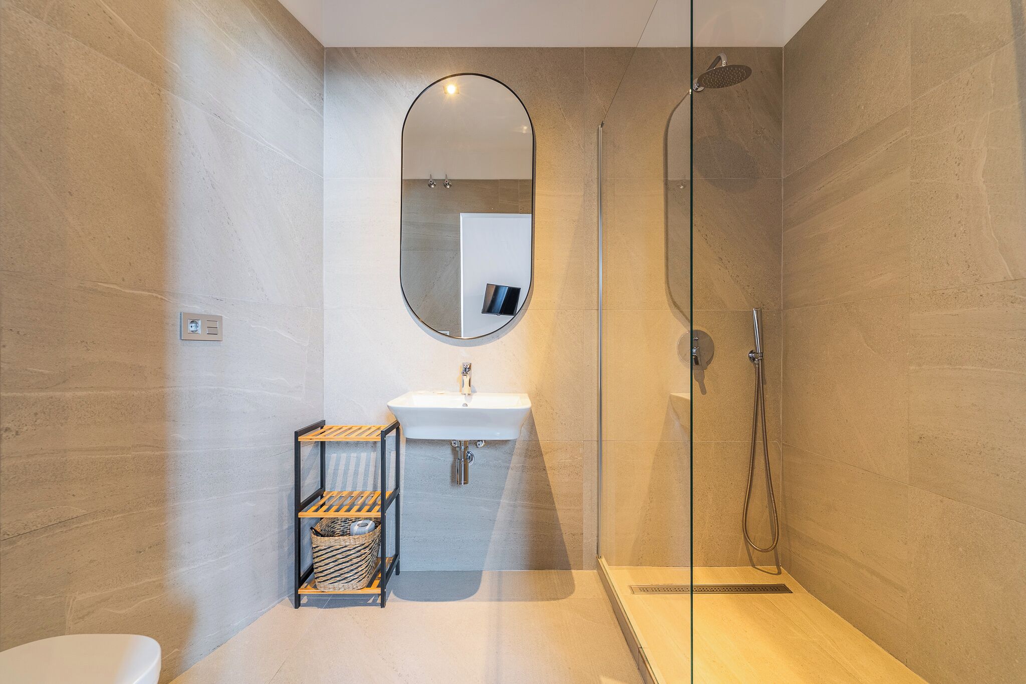 A modern bathroom with a glass shower enclosure, a white sink, and a small wooden shelf with baskets.