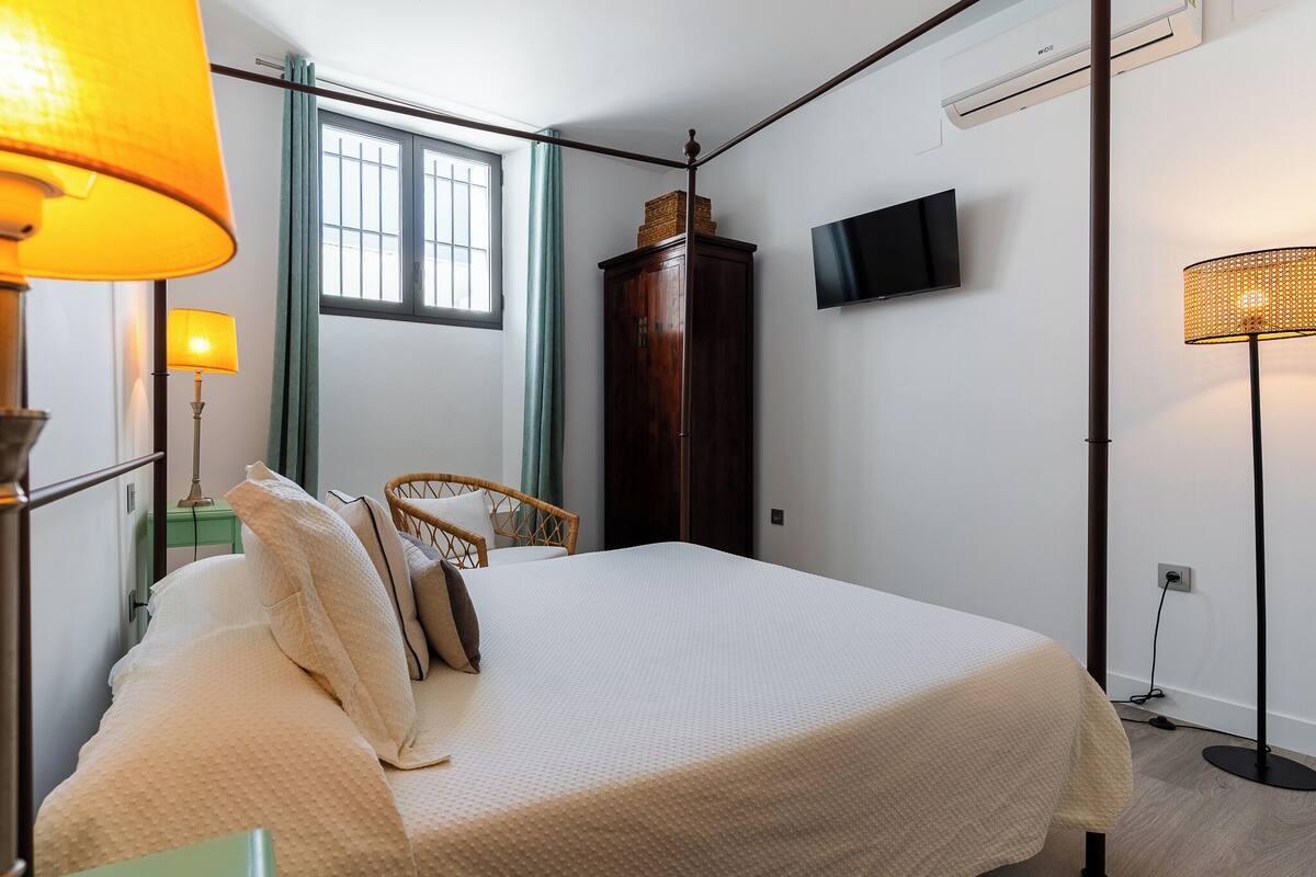 A neatly made bed with a white comforter, a wicker chair, a wooden wardrobe, a television mounted on the wall, and a floor lamp.