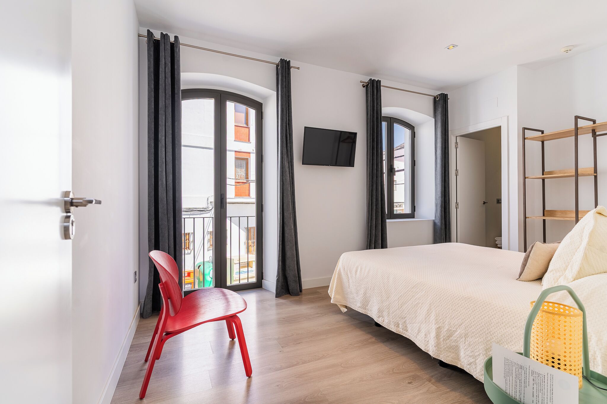 A modern hotel room with a bed, a TV, a balcony with curtains, and a red chair.