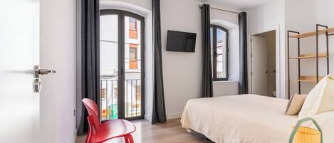 A modern hotel room with a bed, a TV, a balcony with curtains, and a red chair.