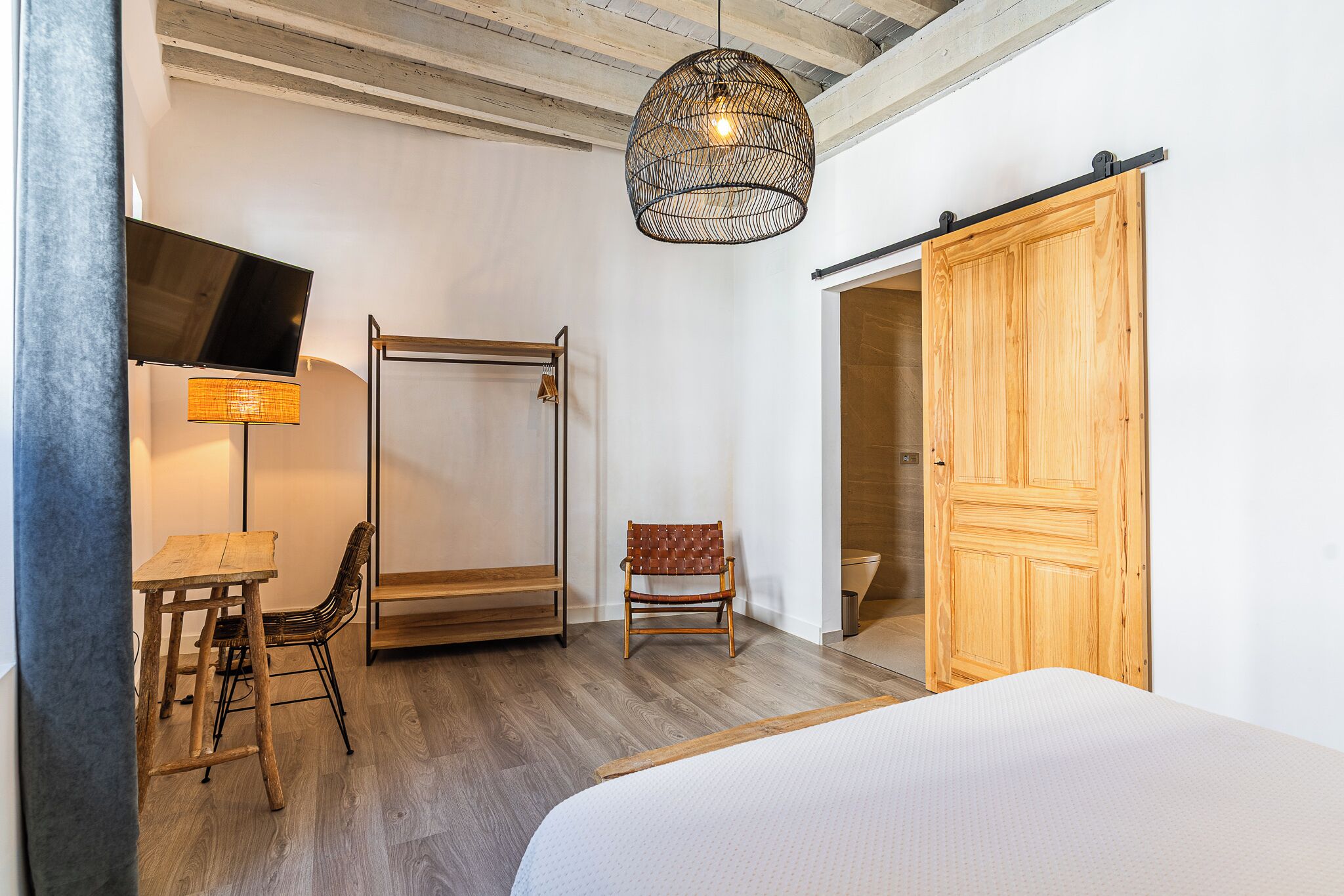 A bedroom with a bed, a desk with a chair, a hanging light fixture, a wooden wardrobe, and a sliding door.