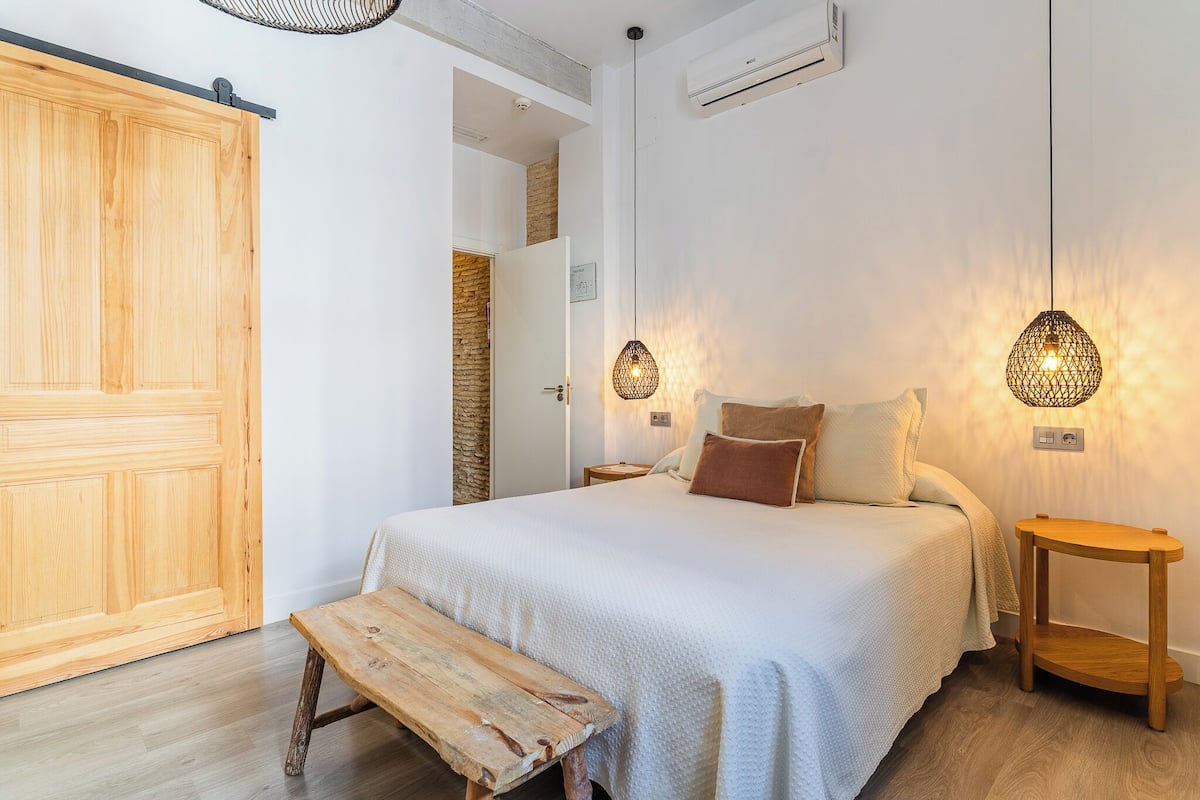 A bedroom with a large wooden wardrobe, a bench, a bed with white bedding, two bedside lamps, and a small wooden nightstand.