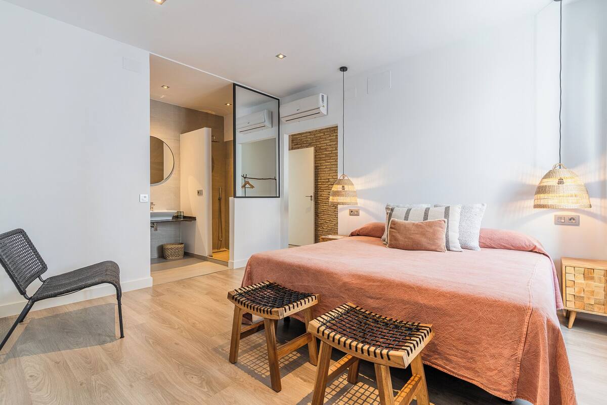 A modern bedroom with a large bed, two wooden stools, a chair, and a bathroom visible through an open door.