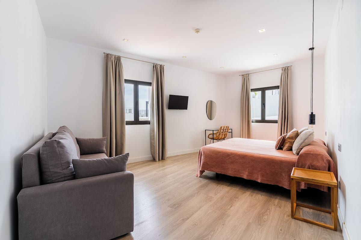 A modern hotel room with a bed, a sofa, a small table, a TV, and large windows with curtains.