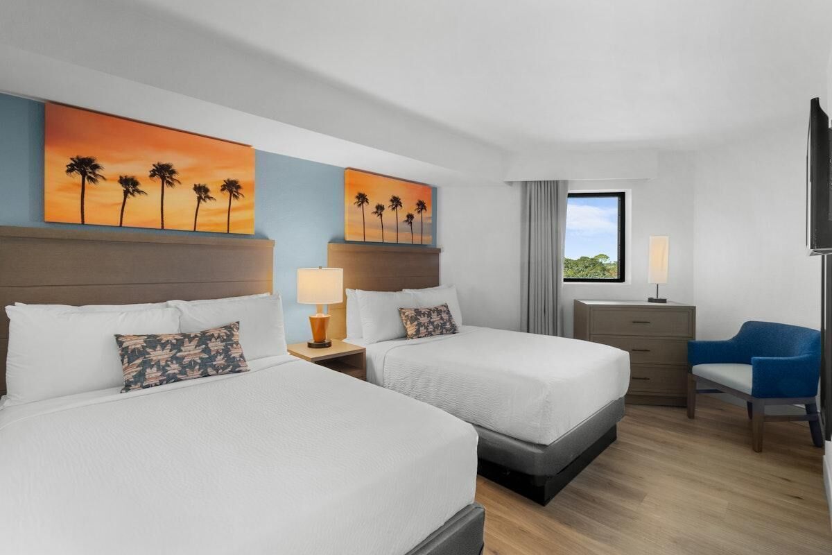 A hotel room with two beds, a desk, a chair, and artwork depicting palm trees.