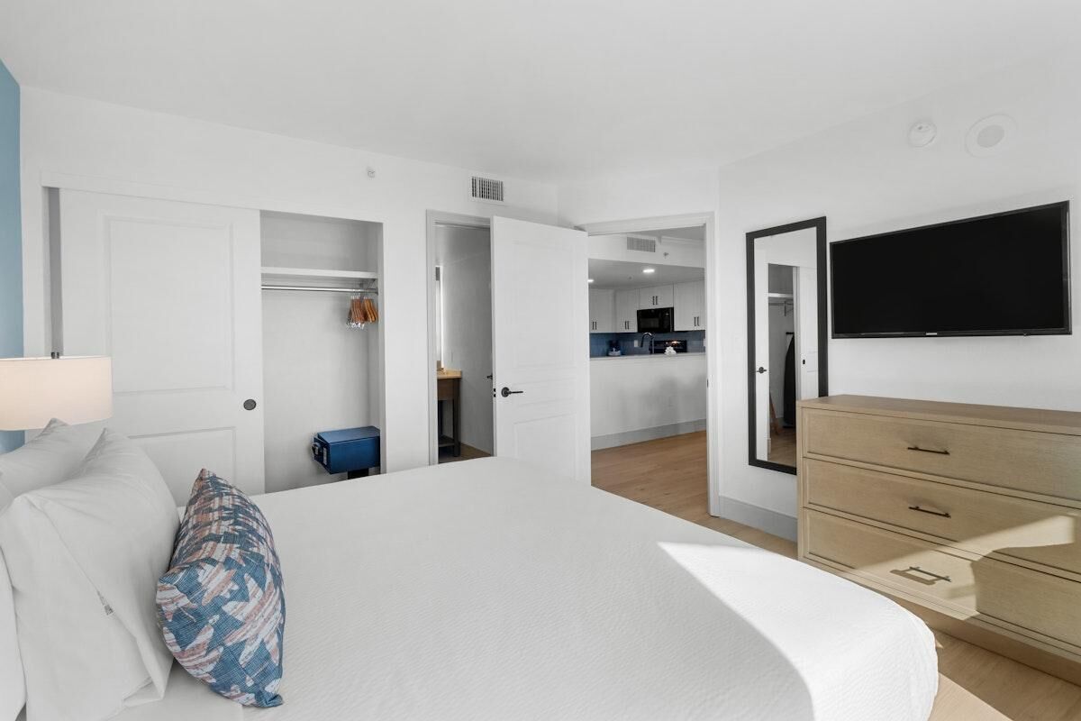 A hotel room with a bed, a dresser, a TV, and a kitchenette in the background.
