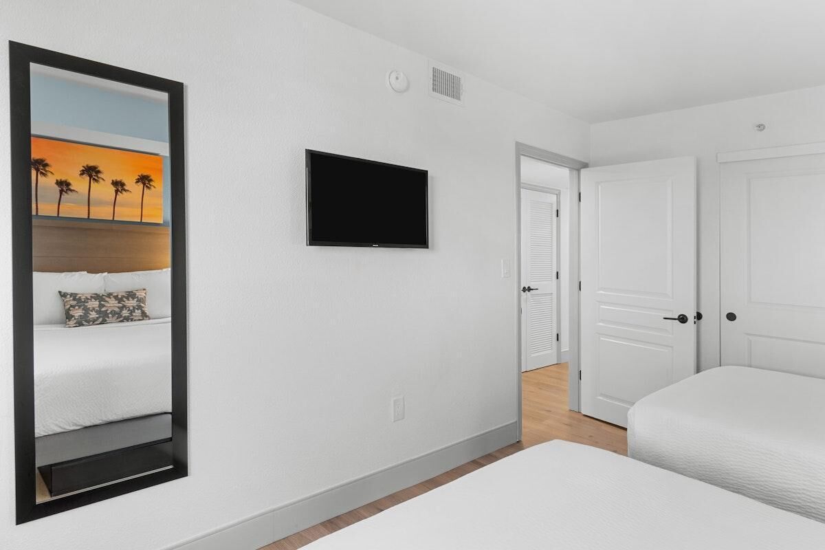 Ein Hotelzimmer mit einem Bett, einem Fernseher, einem großen Spiegel und einer Tür zu einem weiteren Raum.