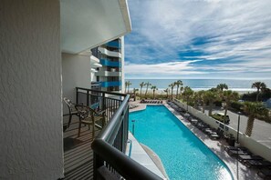 Enjoy the ocean view from this hotel balcony overlooking a rectangular pool with lounge chairs and umbrellas.