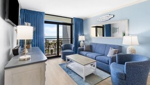 A modern living room with a sofa, armchairs, a coffee table, and a TV. There is a balcony with a view of the beach. - Coastal Getaway 4th Flr 3BR Angle Ocean View Condo (Myrtle Beach)