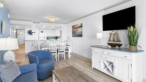 A modern living room with a blue sofa, a wooden coffee table, and a white cabinet with drawers. There is a dining area with white chairs and a table in the background. - Coastal Getaway 4th Flr 3BR Angle Ocean View Condo (Myrtle Beach)