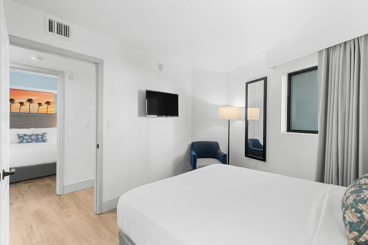 A hotel room with a bed, a TV, a chair, a lamp, and a painting of a sunset.