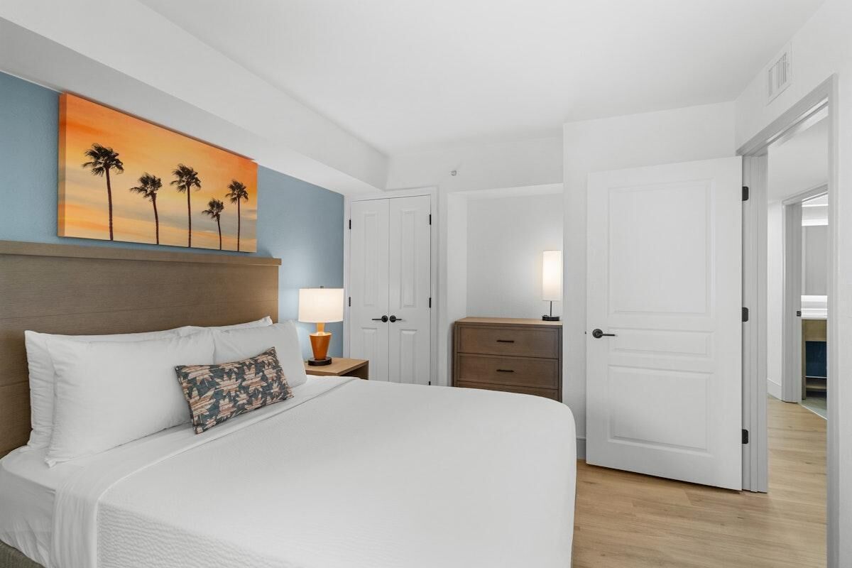 Una camera d'albergo con un letto grande, un comodino con una lampada, un settimino in legno e una porta che dà su un'altra stanza.
