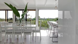 Een moderne eetruimte met witte stoelen en een tafel, grote groene planten en een uitzicht door grote ramen.
