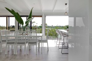 A modern dining area with white chairs and a table, large green plants, and a view through large windows.
