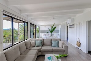 Una sala de estar moderna con un sofá modular, ventanas amplias, un comedor y una cocina al fondo.