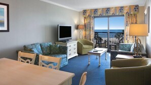 A hotel room with a living area featuring a sofa, chairs, a TV, a dining table, and a balcony with a view.