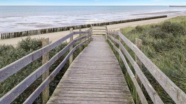 Ein Holzsteg führt zu einem Strand, eine Seite gesäumt von einem Holzzaun, die andere von Grasbewuchs.