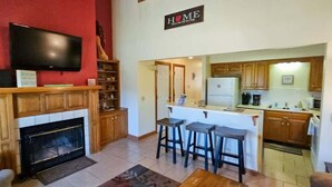 A living room with a fireplace, a TV mounted above it, and a kitchen area with a bar stool. - Great location for exploring Northwest Arkansas! (Holiday Island)