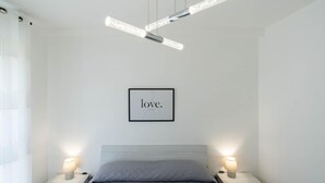 Une chambre à coucher au style minimaliste, avec un lit, deux lampes de chevet, un luminaire au plafond et une citation inspirante encadrée accrochée au mur.