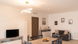 Un salon moderne avec un téléviseur à écran plat, une table à manger avec des chaises, un canapé et une table basse. Des tableaux encadrés ornent le mur, et une porte en bois est visible.
