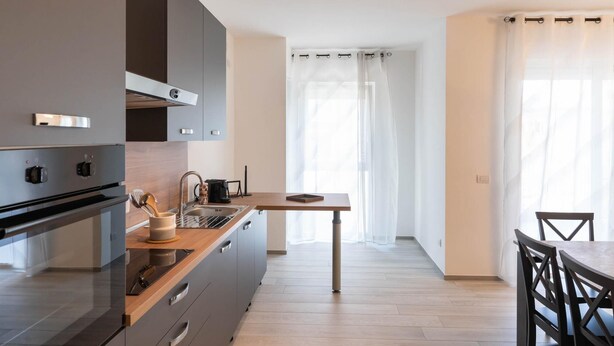 Une cuisine moderne avec des armoires grises, un plan de travail en bois et un four encastré. On y trouve un coin repas avec une table et des chaises, ainsi qu’une fenêtre agrémentée de rideaux légers.
