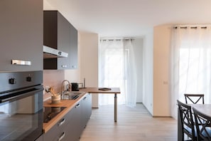 A modern kitchen with grey cabinets, a wooden countertop, and a built-in oven. There is a dining area with a table and chairs, and a window with sheer curtains. - Dimora Rosselli - Appartamento 5 (Fasano)