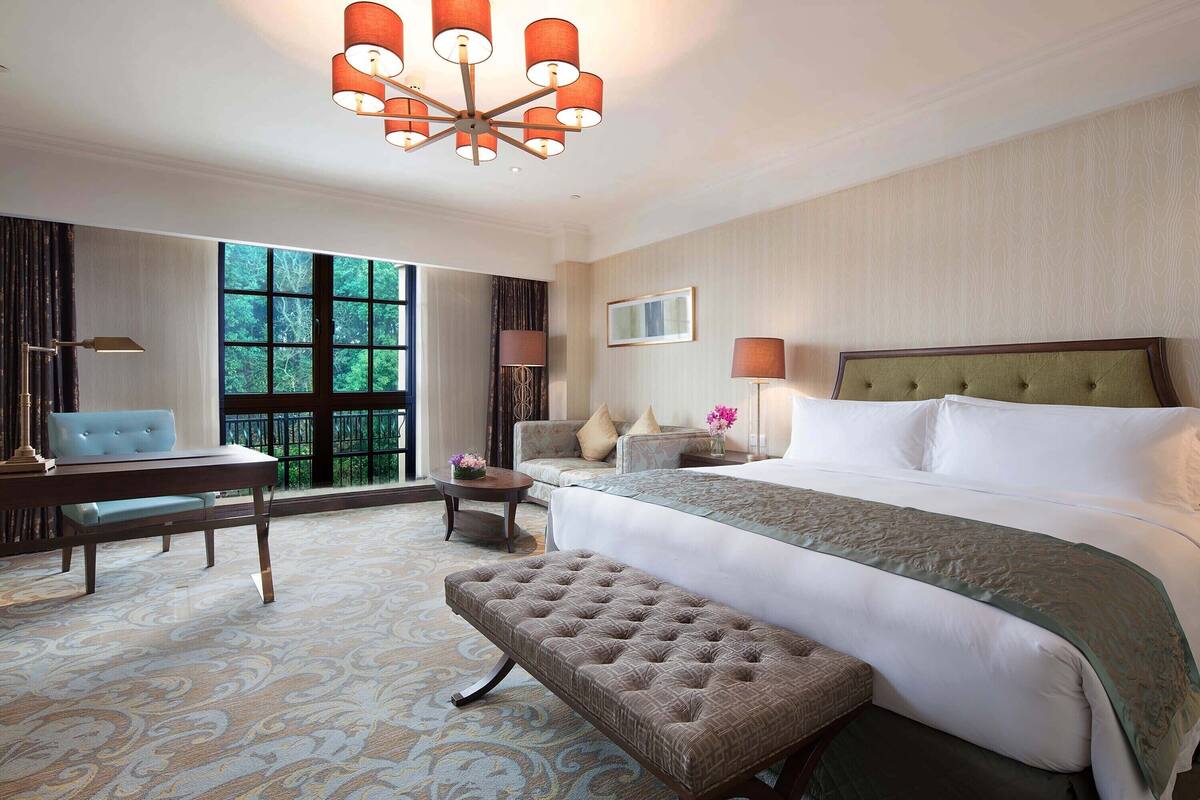 Hotelkamer met een groot bed, een zithoek met een bank en een tafel, een bankje en een groot raam met uitzicht op bomen.