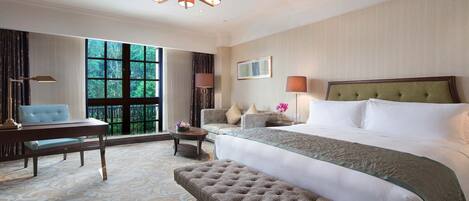 A hotel room with a large bed, a sitting area with a sofa and a table, a bench, and a large window with a view of trees.