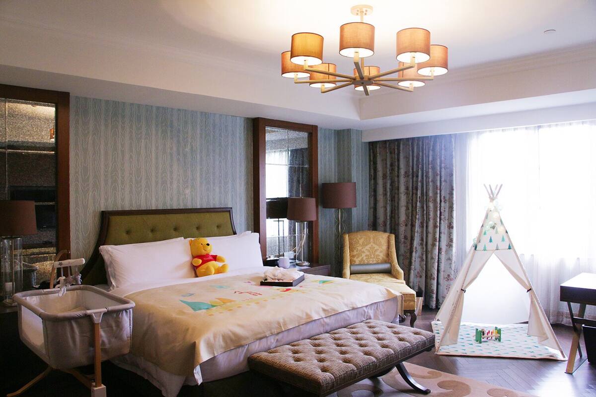 Een hotelkamer met een groot bed, een nachtkastje, een stoel, een lamp, een spiegel, een raam met gordijnen, en een tipi decoratie.