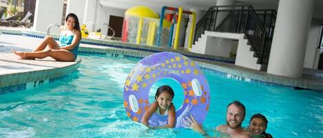 Piscina com uma criança e um adulto na água, área de lazer com espreguiçadeiras e um espaço coberto com escadas e corrimãos.